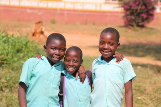 Three smiling schoolchildren in light blue uniforms stand arm-in-arm outdoors, posing for a photo. They appear happy and confident, with a school building and plants in the background under bright sunlight.